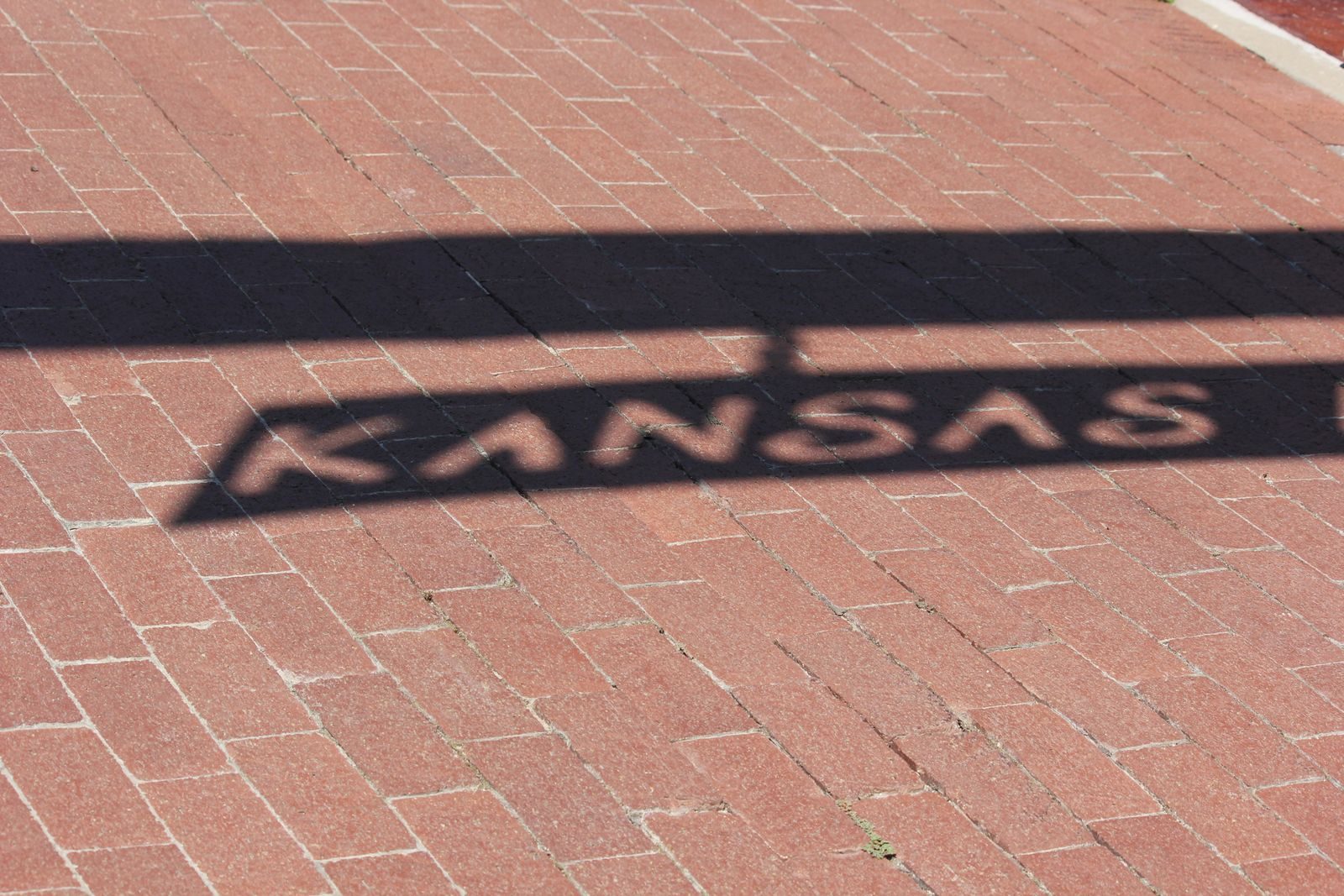 A shadow with the word Kansas stretches across red brick pavement, capturing light and texture.